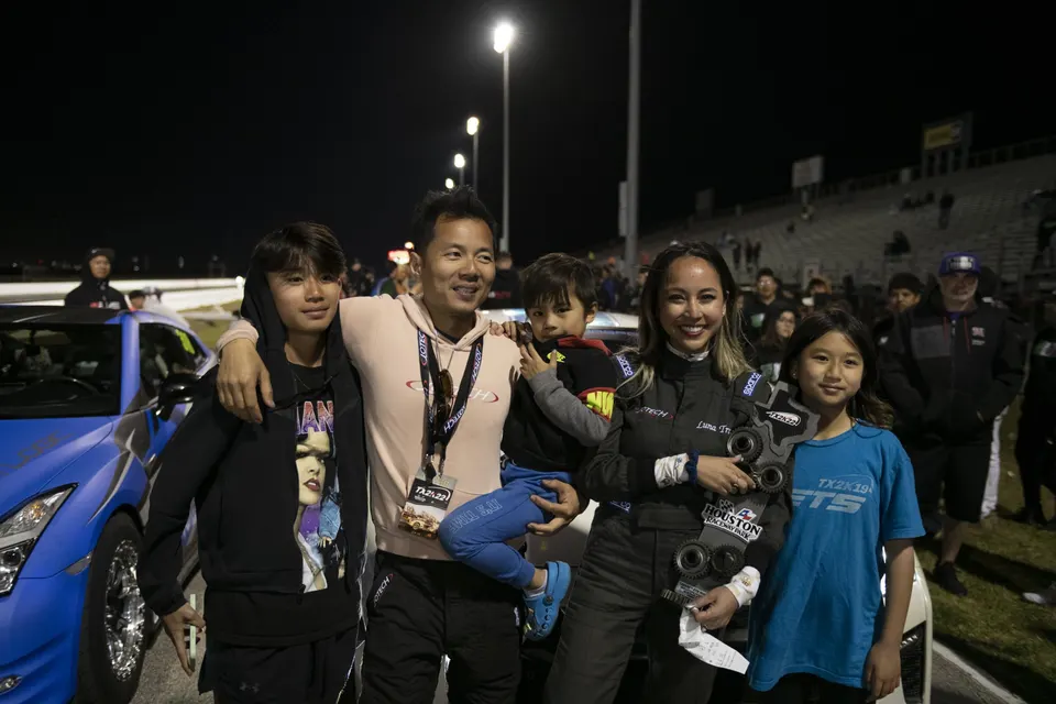 A family celebrates at a night drag race event, standing in front of a blue Nissan GT-R, holding a trophy.