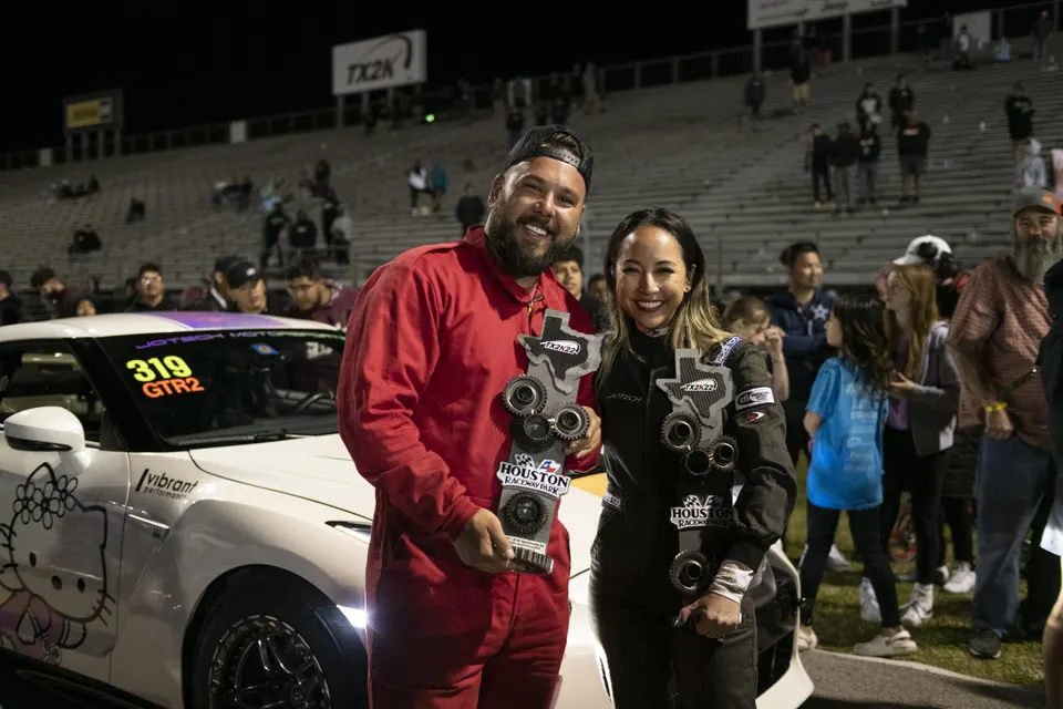 A man and a woman smiling and holding TX2K22 trophies next to a white Nissan GT-R with Hello Kitty graphics at Houston Raceway Park.