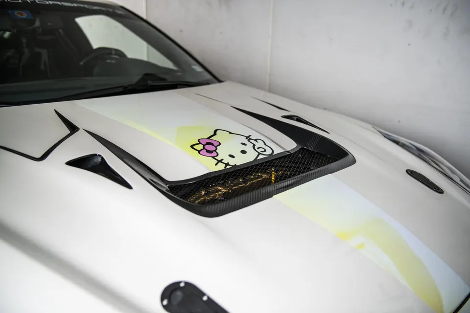 A close-up of a white car's hood featuring carbon fiber vents, a yellow stripe, and a Hello Kitty sticker.