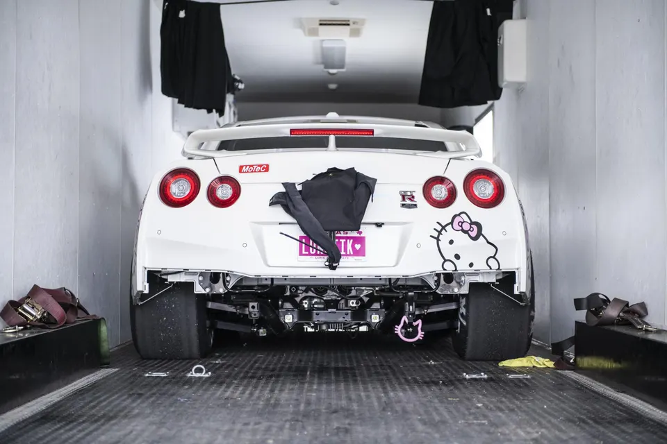 Rear view of a white Nissan GT-R drag car with Hello Kitty decal in a trailer, showcasing exposed suspension and wide drag tires.