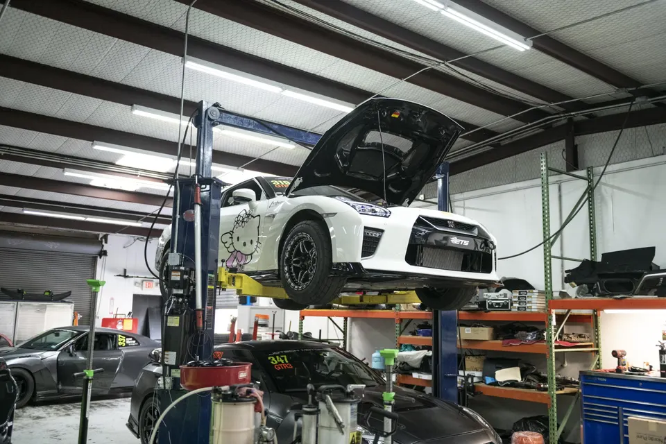 A white Nissan GT-R with a Hello Kitty decal on a car lift in a performance tuning shop with its hood open.