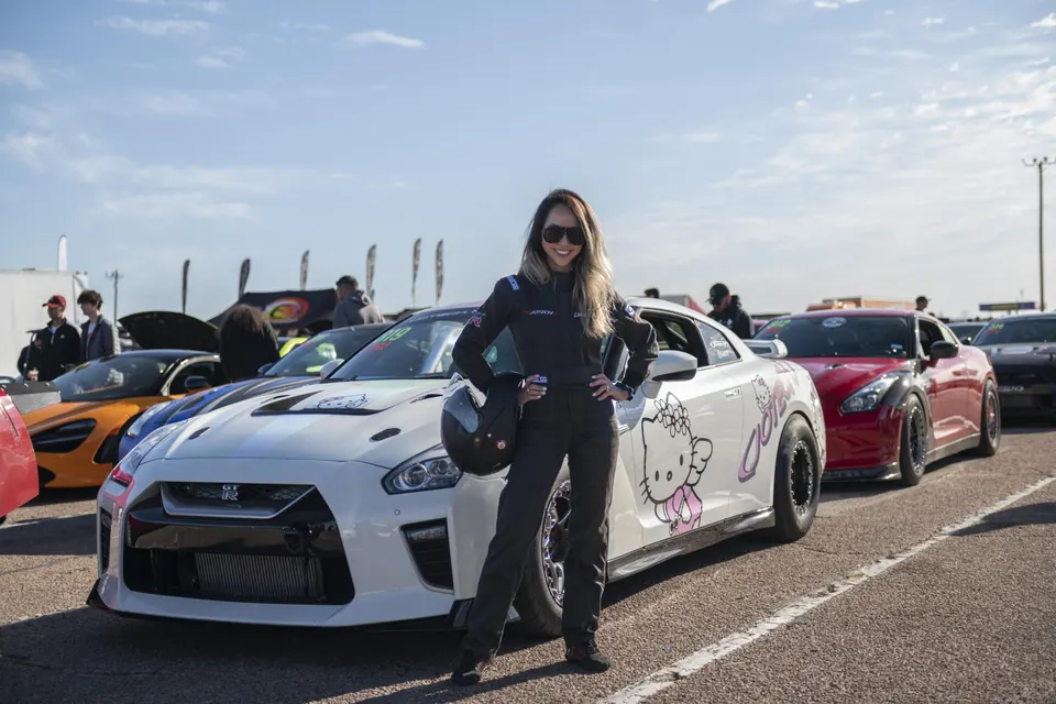 A female driver in a racing suit stands proudly in front of her white Nissan GT-R with Hello Kitty decals at a car event.