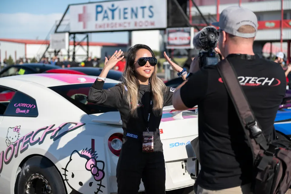 A woman in a racing suit being interviewed next to a white Jotech Nissan GT-R with Hello Kitty decals at a track event.