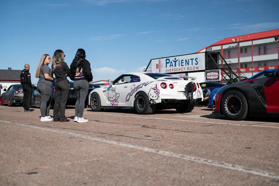 A white Nissan GT-R with a Hello Kitty wrap and drag slicks in a staging lane at a drag strip with other race cars and people.