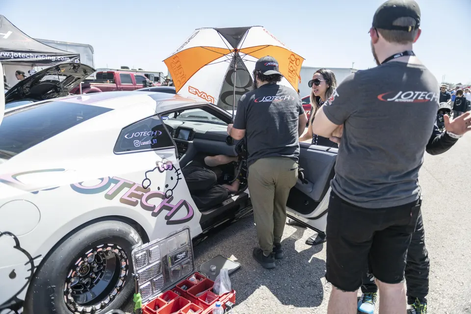 A white Jotech Motorsports Nissan GT-R with Hello Kitty decals being prepped trackside by mechanics at a drag racing event.