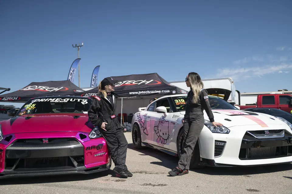 Two women in racing suits lean on a pink Nissan GT-R and a white Hello Kitty Nissan GT-R at a track event.