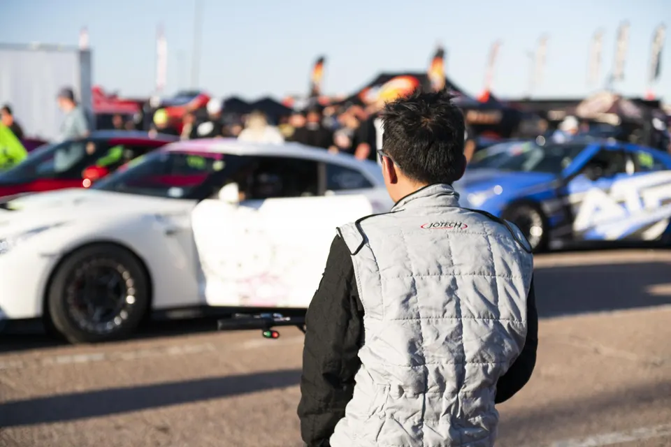 A racer in a Jotech suit stands at a track event, looking at a white Nissan GT-R and other performance cars.