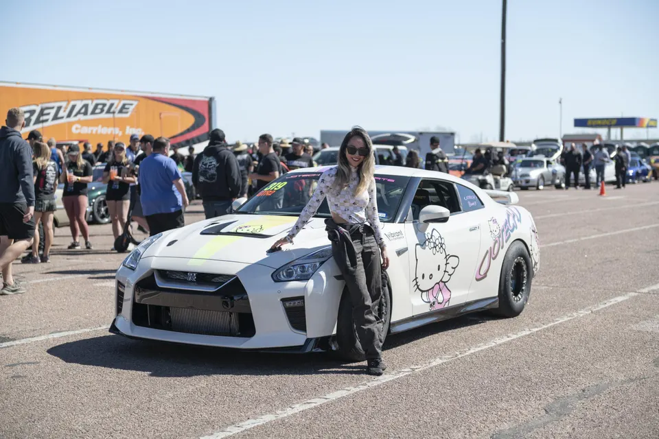 A white Nissan GT-R with Hello Kitty livery and a woman posing next to it at a drag strip event.