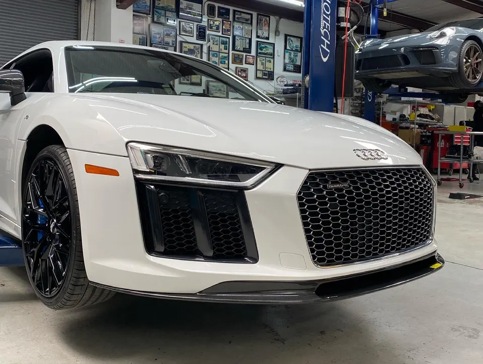 White Audi R8 on a lift in a performance shop, showcasing its front grille and carbon fiber splitter.