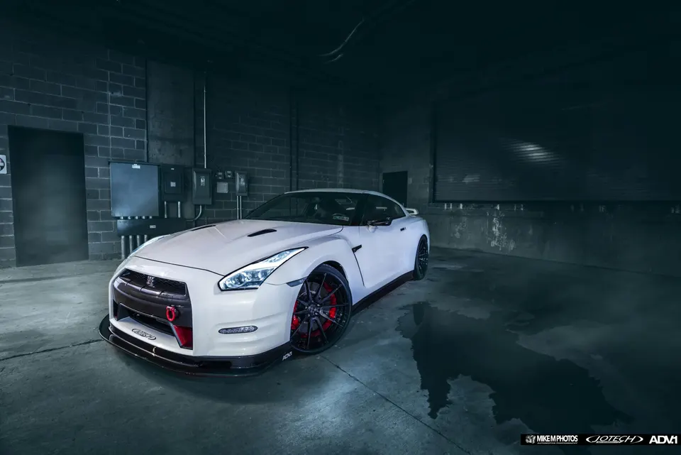 A white Nissan GT-R with aftermarket wheels and a front splitter parked in a dimly lit garage.
