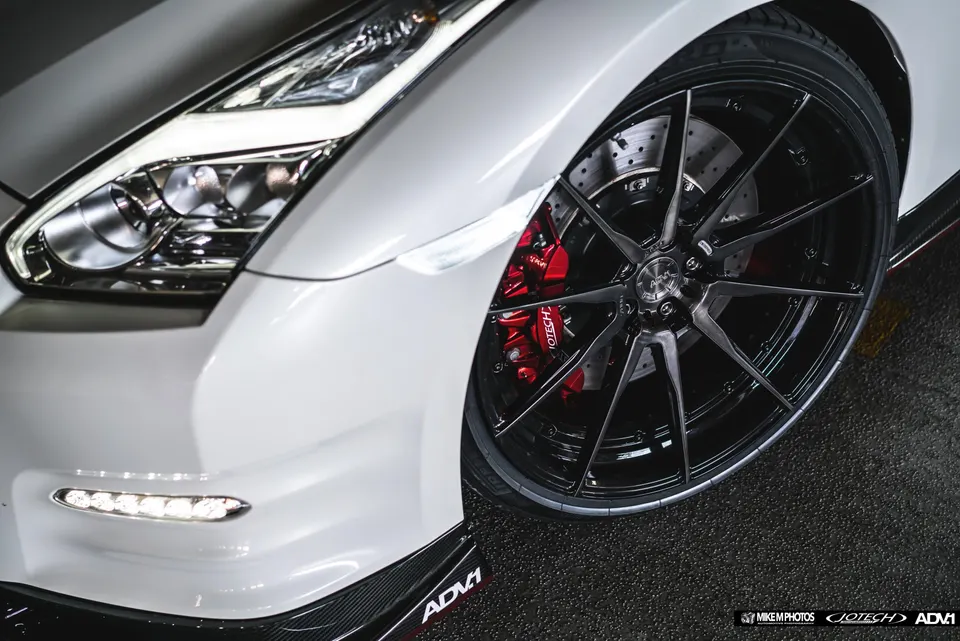 Close-up of a white Nissan GT-R's front wheel, featuring ADV.1 wheels, red brake calipers, and a carbon fiber splitter.
