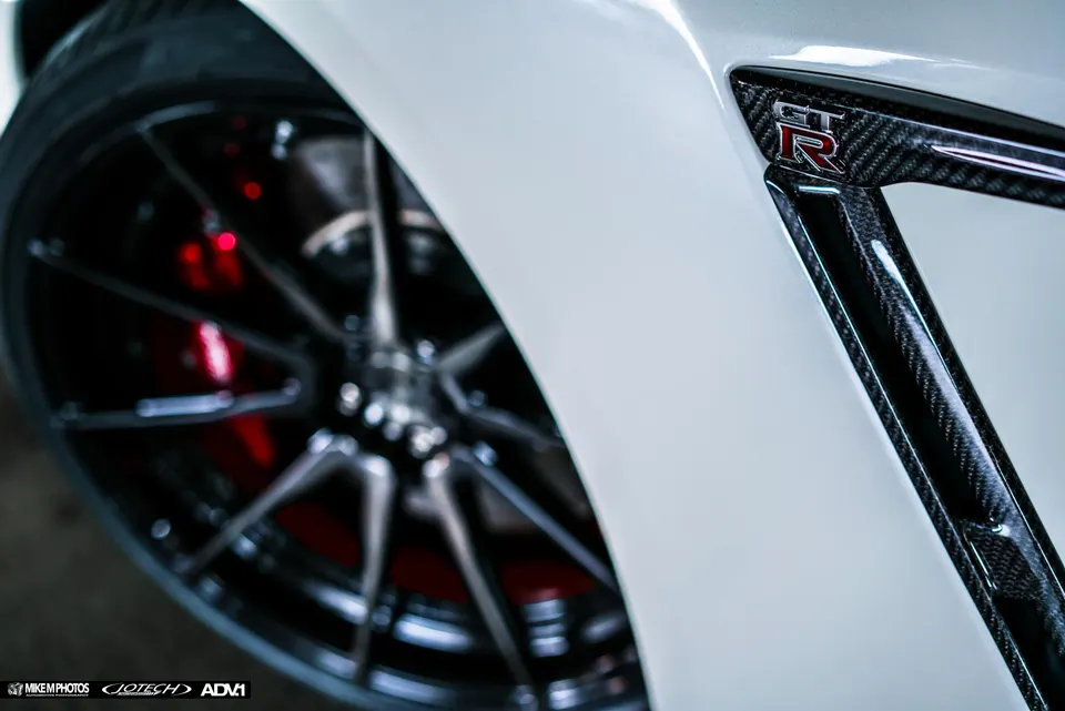 Close-up of a white Nissan GT-R's front fender with carbon fiber trim, ADV.1 wheel, and red brake calipers.