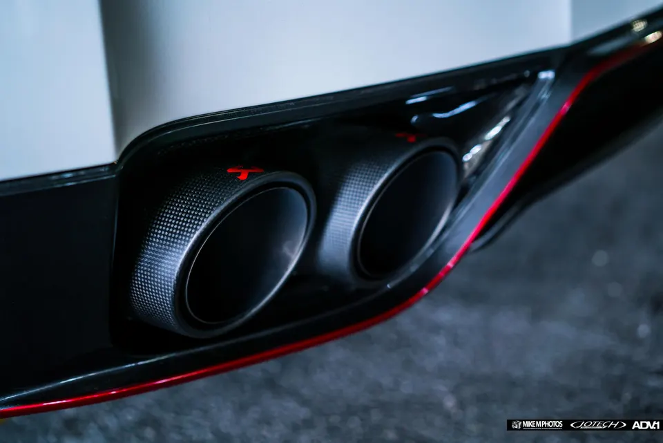 Close-up of carbon fiber exhaust tips with red accents on a performance car's rear bumper, showcasing intricate detail.