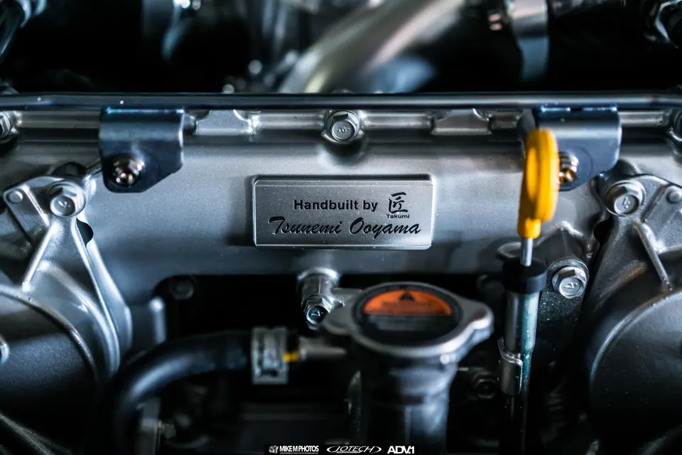 A close-up of a handbuilt engine block with a 'Tsuneomi Ooyama' plaque in a performance car's engine bay.