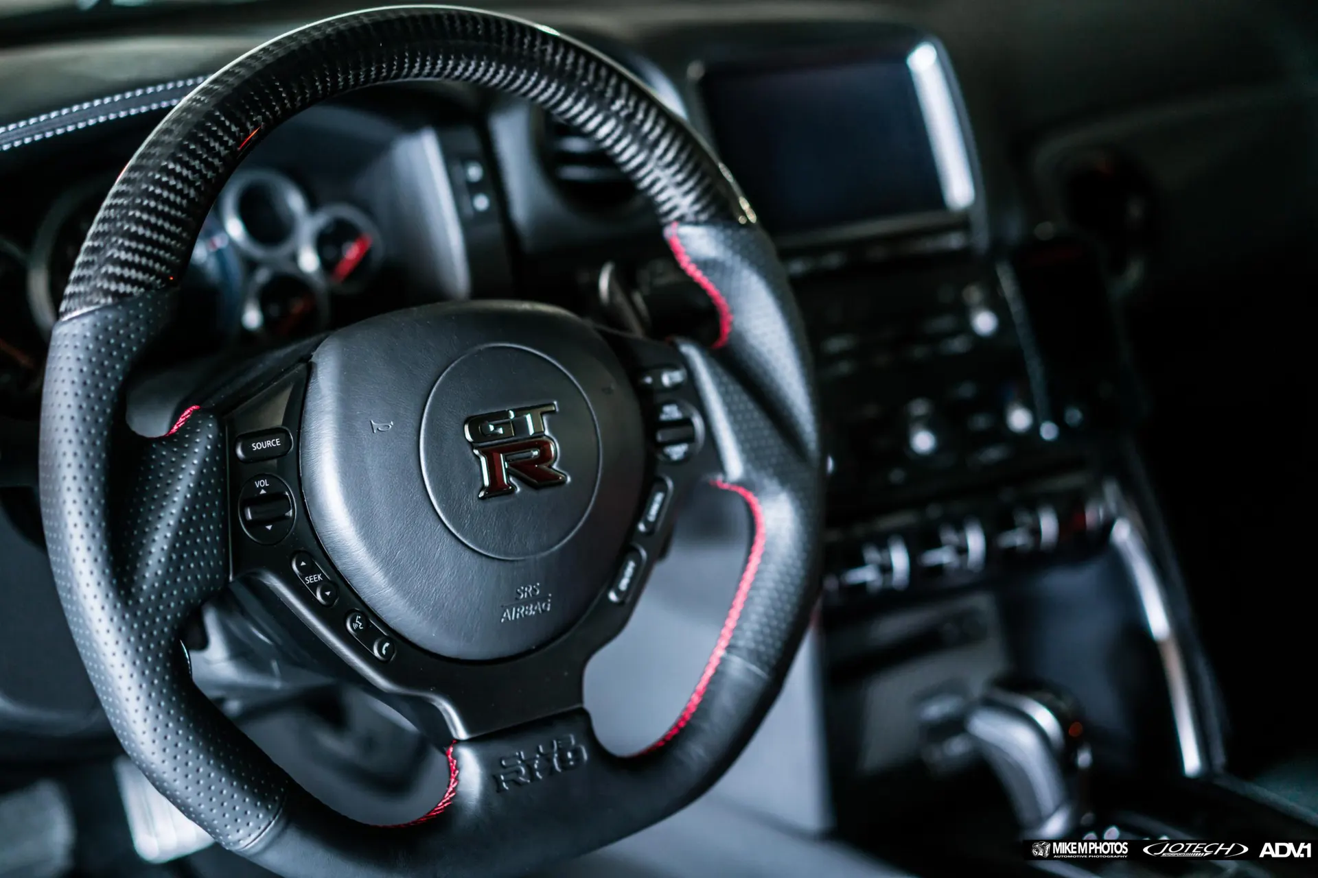 Close-up of a Nissan GT-R's carbon fiber steering wheel with red stitching and interior details.