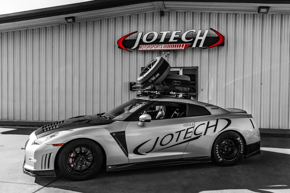 Silver Nissan GT-R with Jotech Motorsports livery and roof rack carrying a race tire, parked outside the Jotech shop.