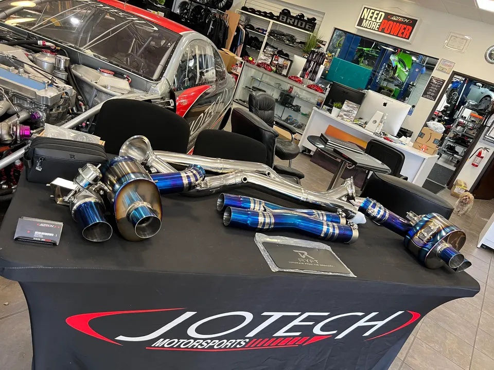 Aftermarket titanium exhaust system components displayed on a Jotech Motorsports table in a performance tuning shop with a race car in the background.