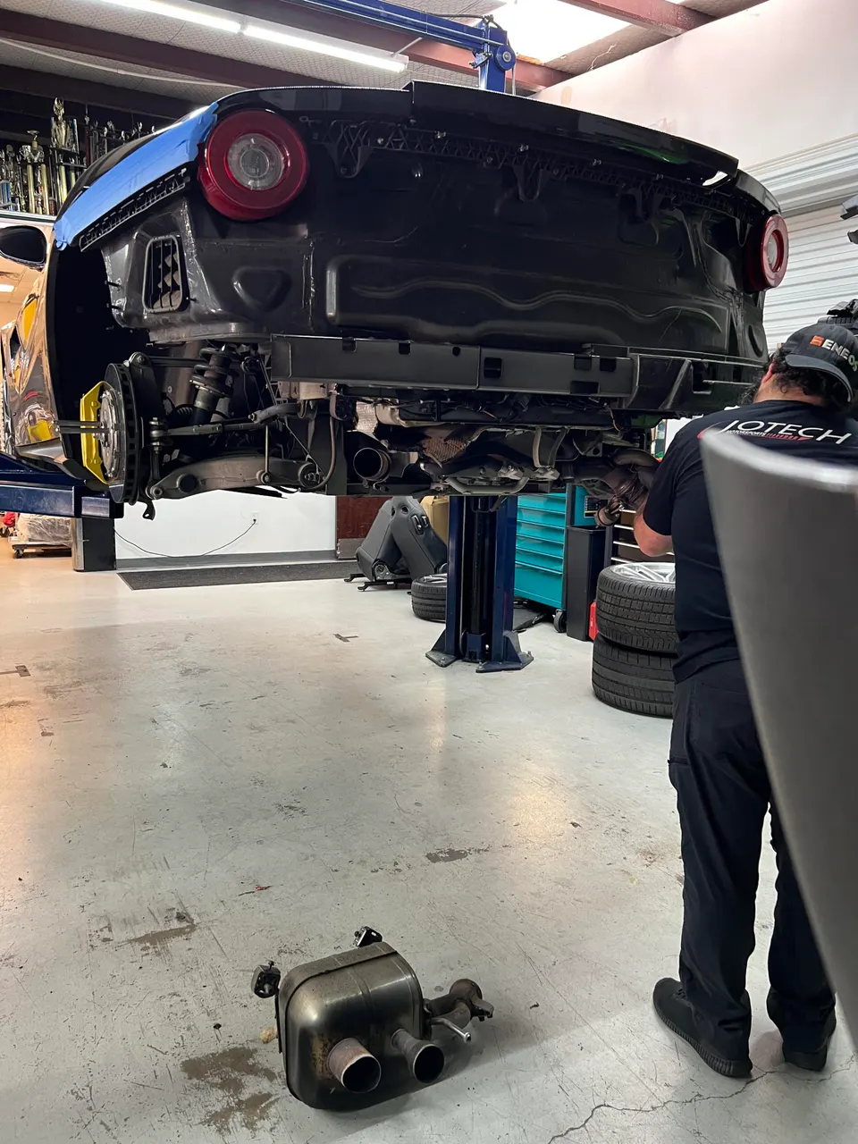 A black Ferrari on a lift in a Jotech Motorsports shop with its rear bumper removed, a mechanic working on the exhaust system.
