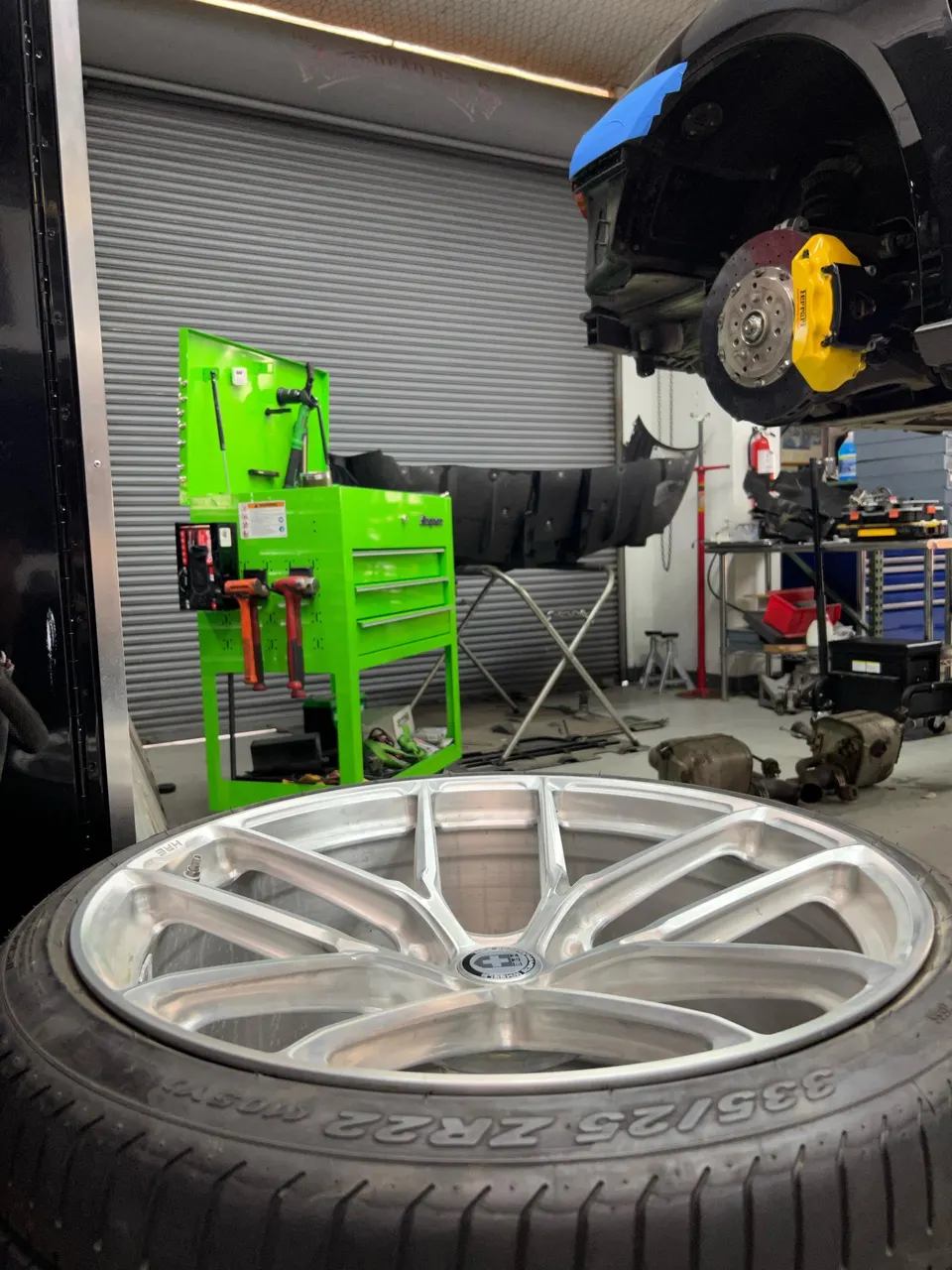 A silver HRE wheel and tire in the foreground of a Jotech Motorsports shop, with a car on a lift showing yellow brake calipers.