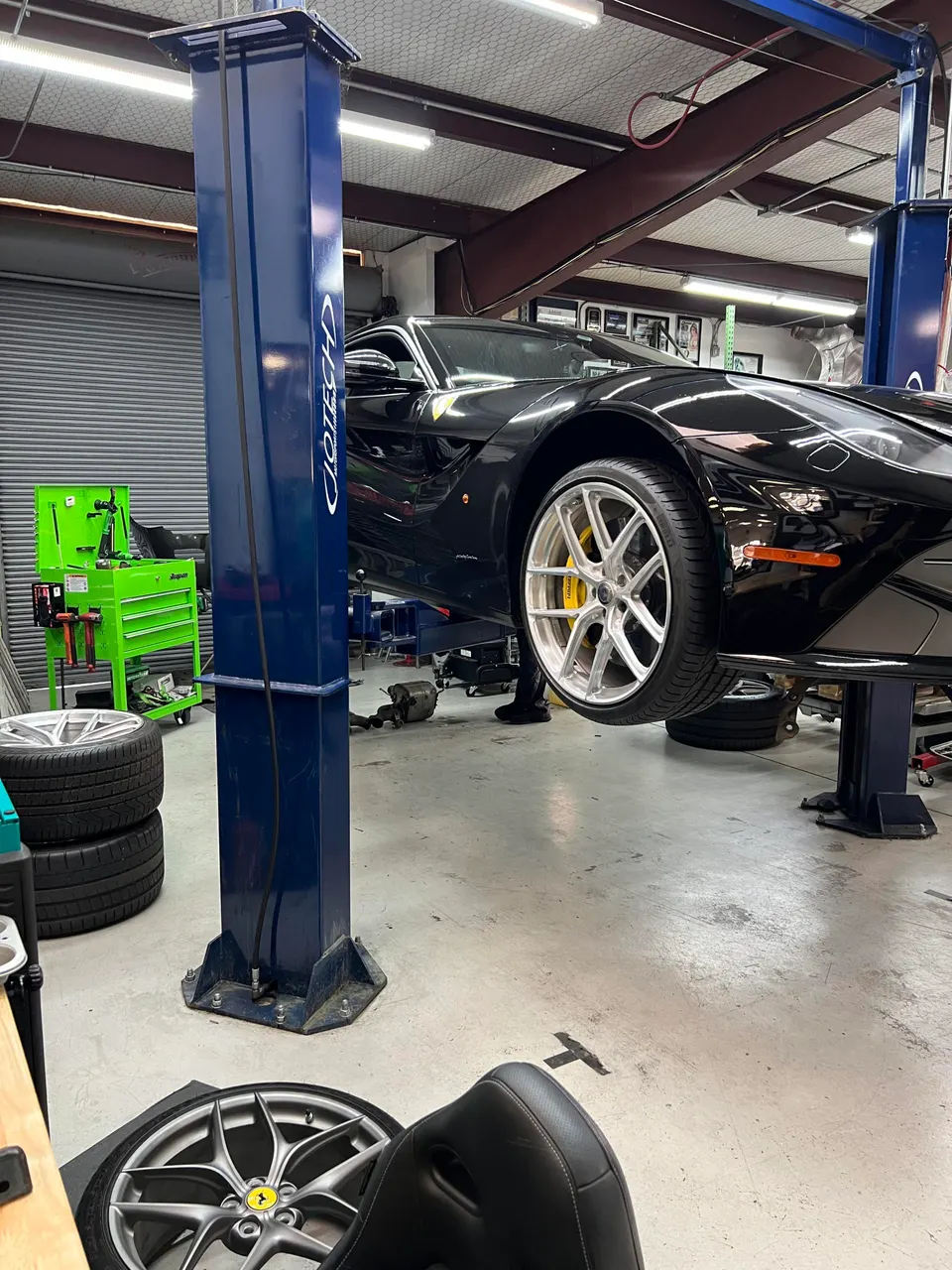 A black Ferrari F12 on a lift in a Jotech Motorsports shop with aftermarket wheels and yellow brake calipers.