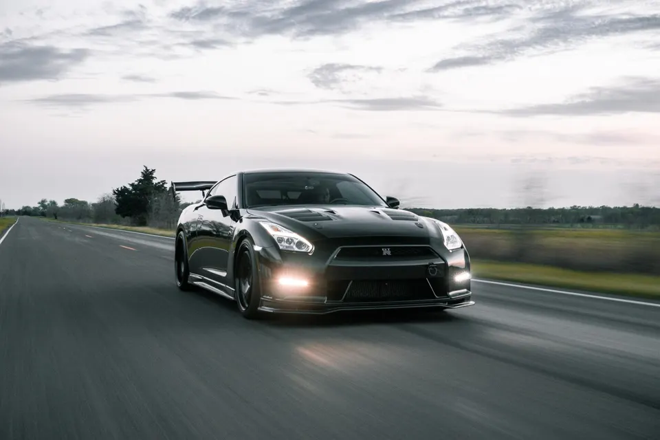 A black Nissan GT-R with an aftermarket wing drives on a road at dusk, showcasing its sleek design.