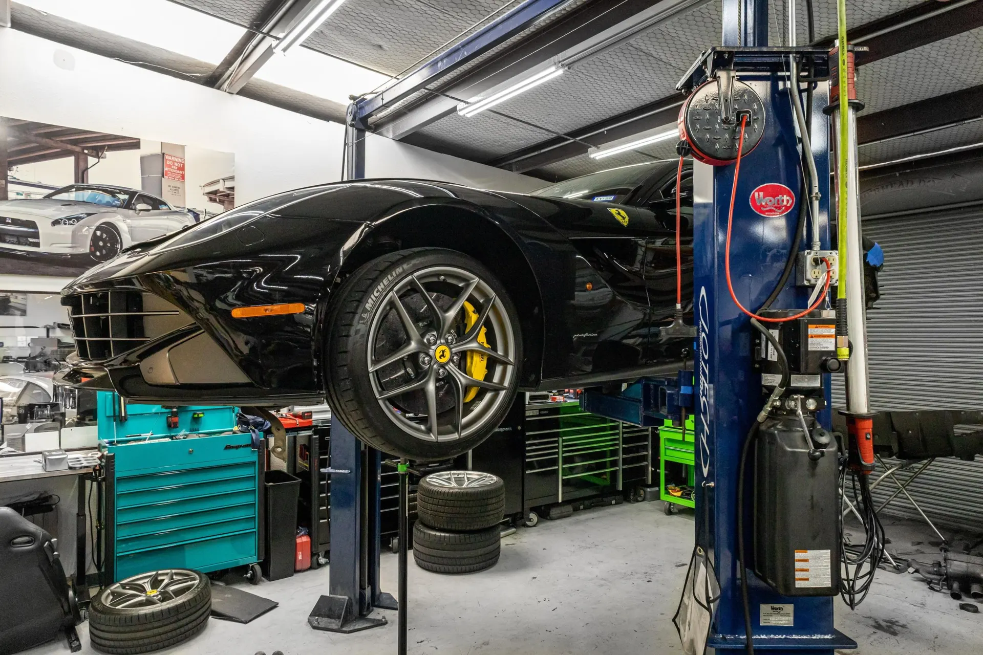 A black Ferrari F12berlinetta on a car lift in a performance tuning shop, with tools and spare wheels visible.