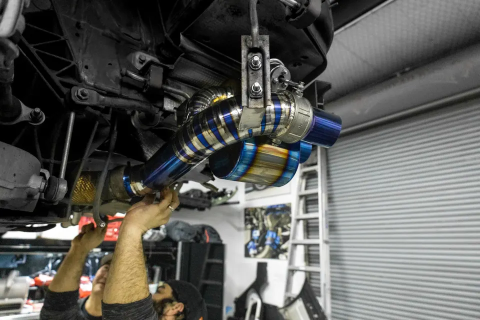Mechanics installing a vibrant blue and gold titanium aftermarket exhaust system on a car in a performance shop.
