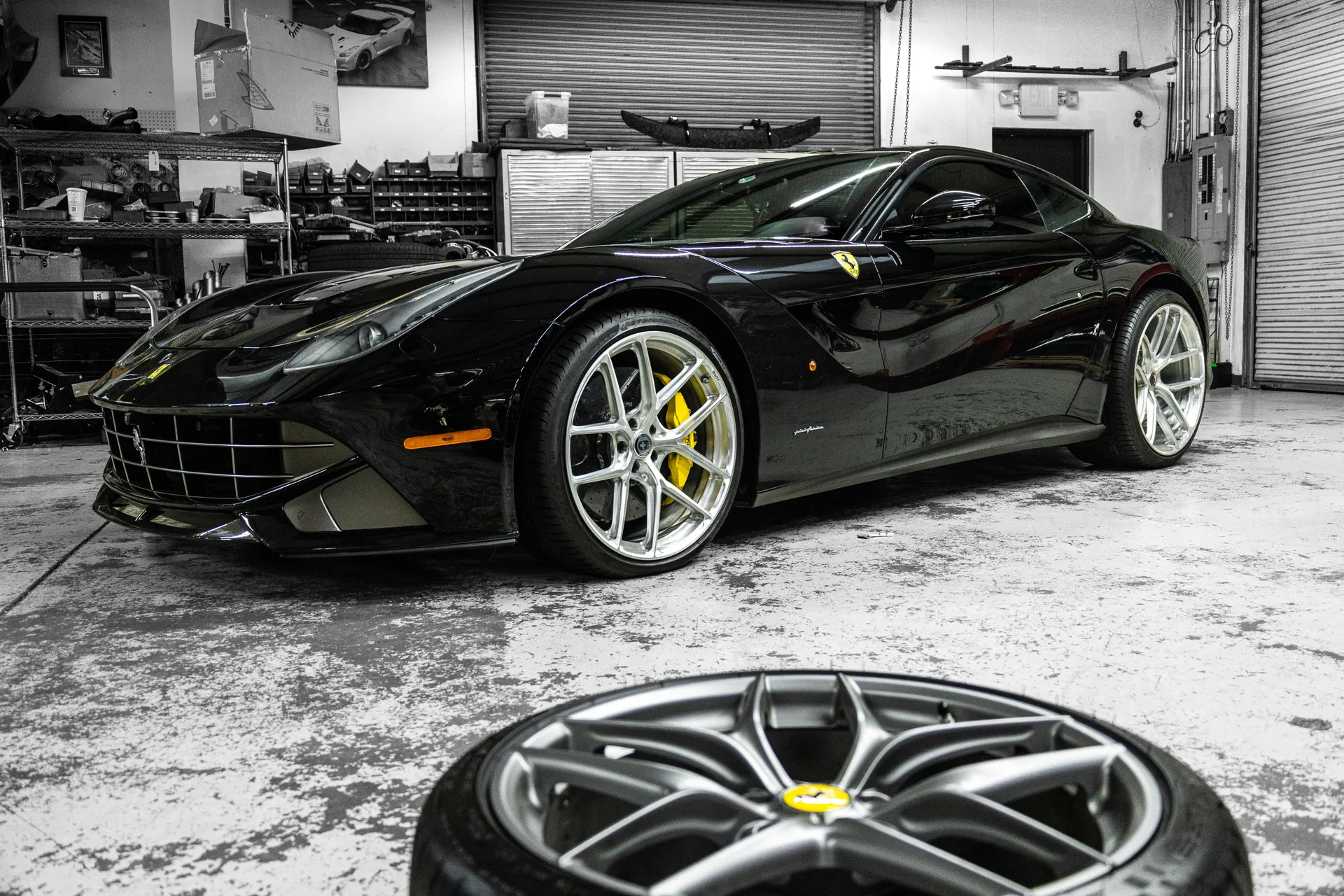 A black Ferrari F12berlinetta with custom silver wheels and yellow brake calipers in a performance shop.