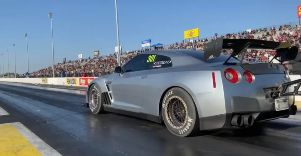 A silver Nissan GT-R drag car with Hoosier slicks and a large rear wing on a drag strip with spectators.