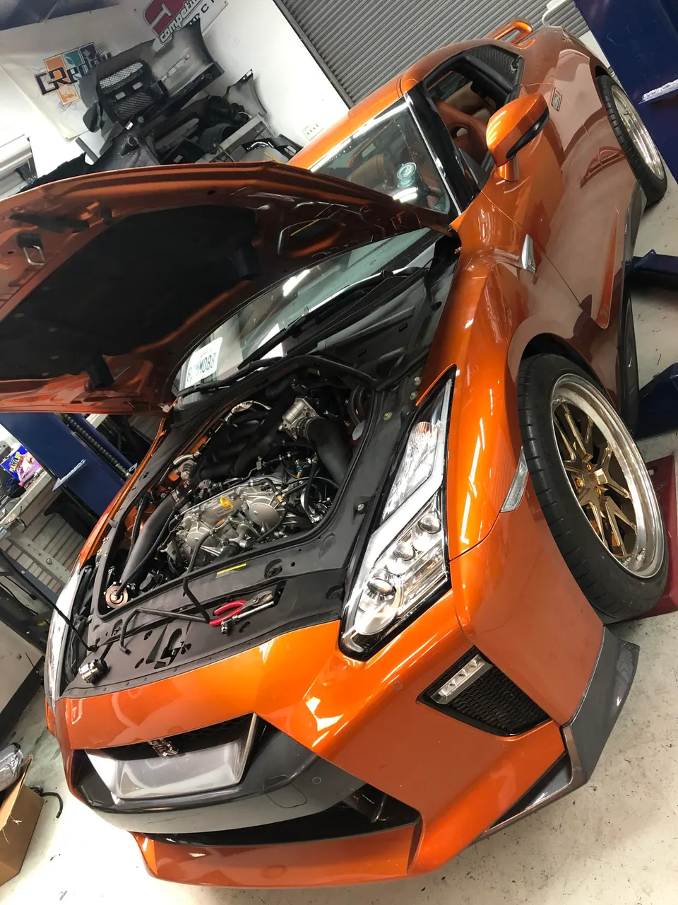 Orange Nissan GT-R with its hood open, revealing the engine bay and induction system, in a Jotech Motorsports shop.