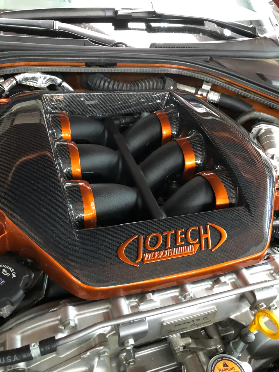 Close-up of a custom carbon fiber intake manifold with orange accents in a high-performance engine bay, featuring Jotech Motorsports branding.