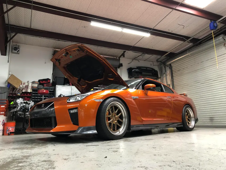 An orange Nissan GT-R with its hood open, parked in a performance tuning shop with tools and parts in the background.