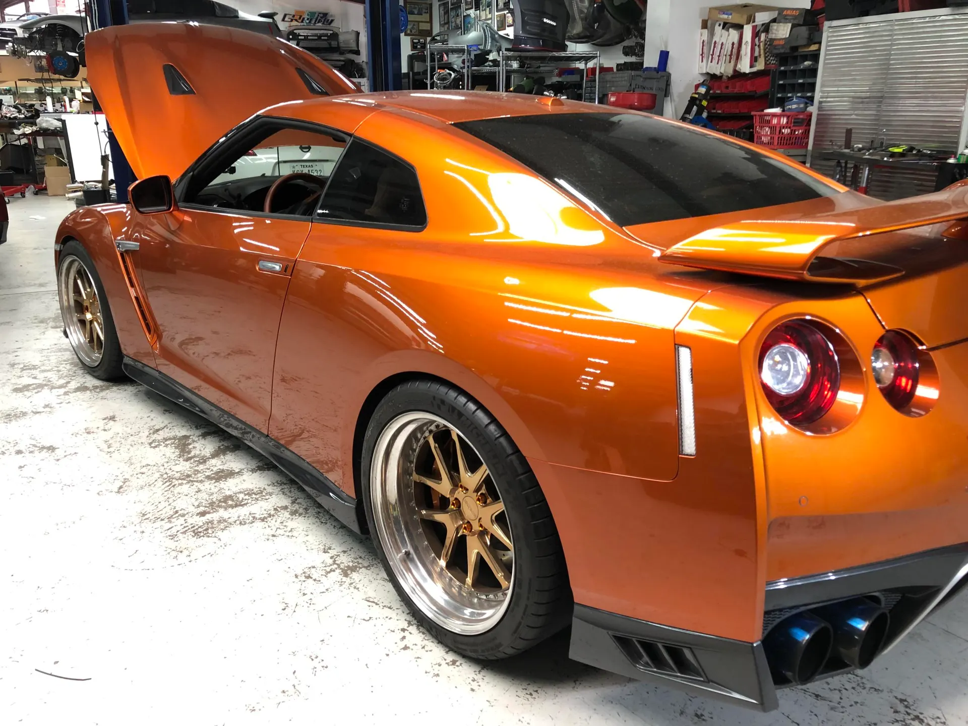 An orange Nissan GT-R with its hood open and aftermarket wheels sits in a performance tuning shop.