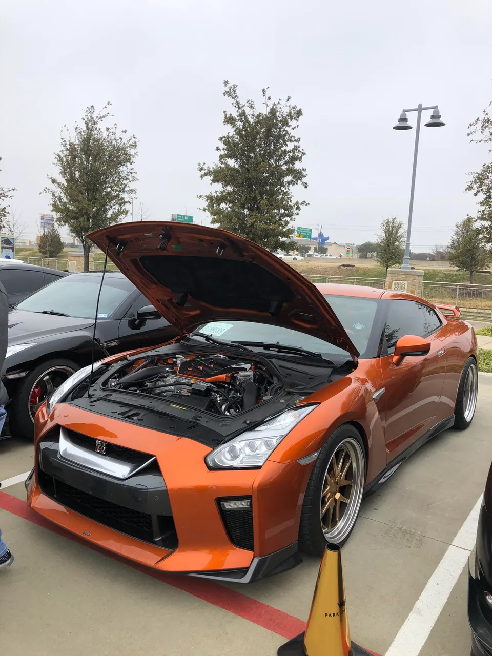 Orange Nissan GT-R with its hood open, showcasing a modified engine bay and aftermarket wheels at an outdoor car meet.