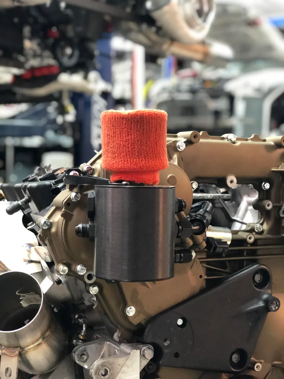 A close-up of a performance engine with an orange air filter and a black catch can in a shop environment.