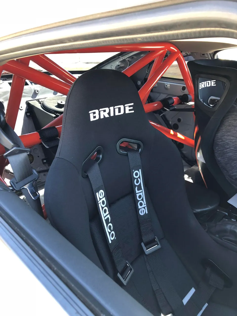 Close-up of a black Bride racing seat with Sparco harnesses and a red roll cage inside a performance car.