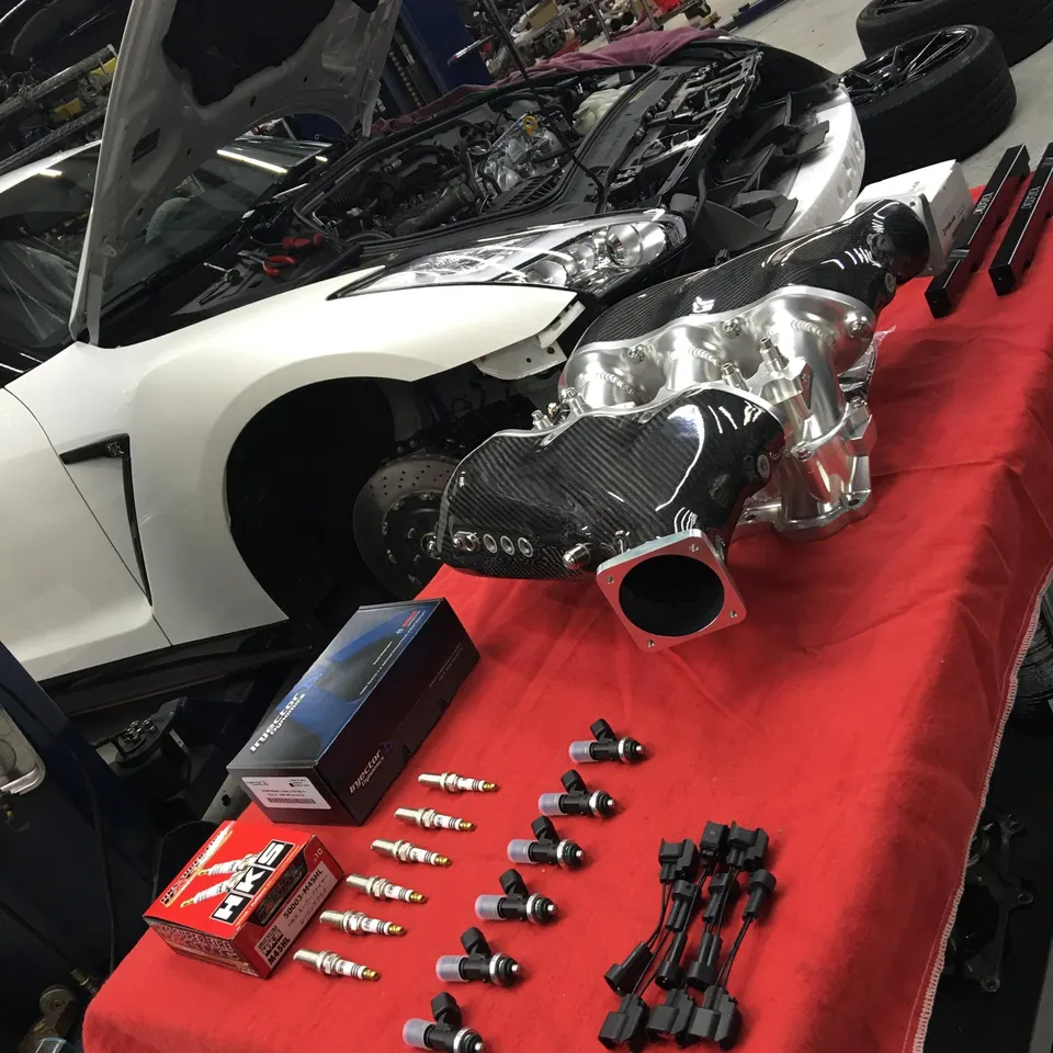 White Nissan GT-R in a shop with its hood open, surrounded by performance parts including a carbon fiber intake manifold, fuel injectors, and spark plugs.