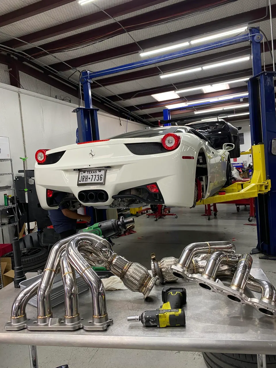 A white Ferrari 458 on a lift in a shop with performance exhaust headers and tools on a workbench.