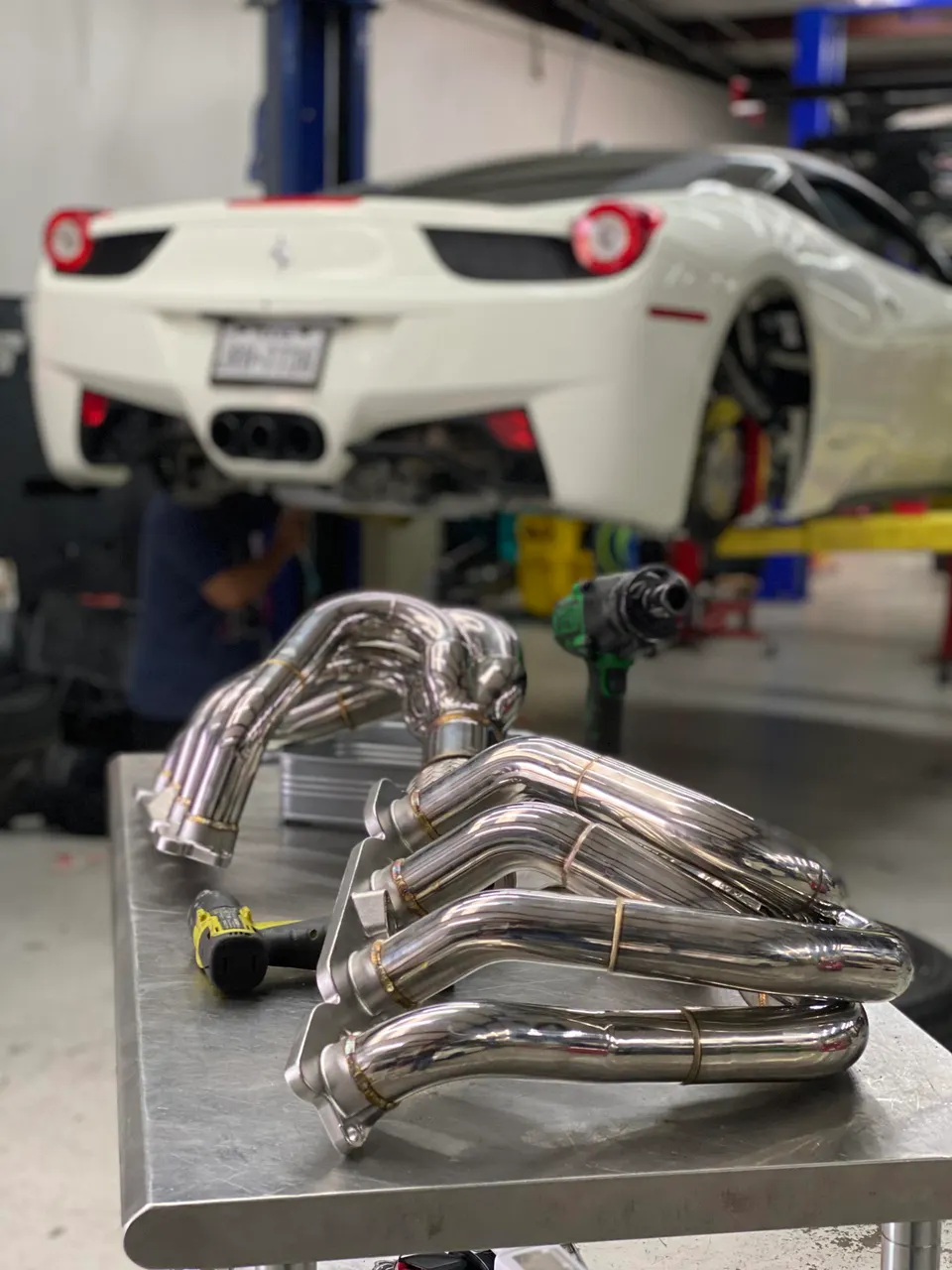 White Ferrari 458 on a lift in a shop with new performance exhaust headers on a workbench.