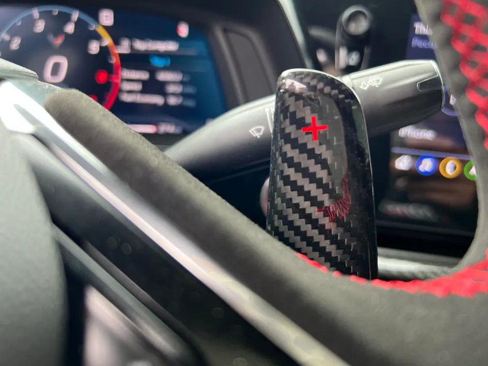Close-up of a carbon fiber paddle shifter with red plus sign and red stitching on a Corvette steering wheel.