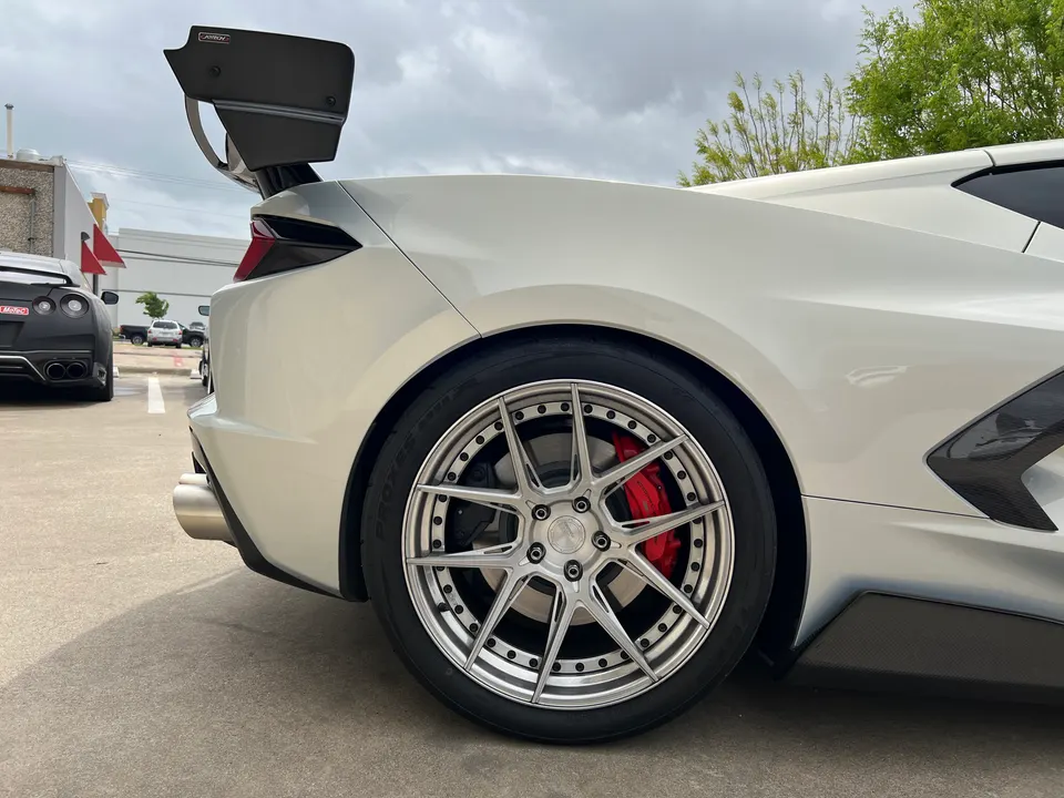 A white Corvette C8 with aftermarket multi-spoke wheels, red brake calipers, and a large carbon fiber rear wing.