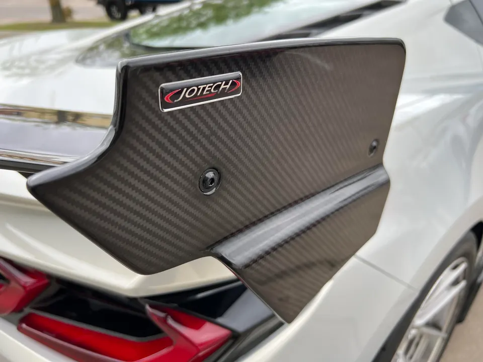 A close-up of a Jotech carbon fiber rear wing on a white Corvette, showcasing the weave and logo.