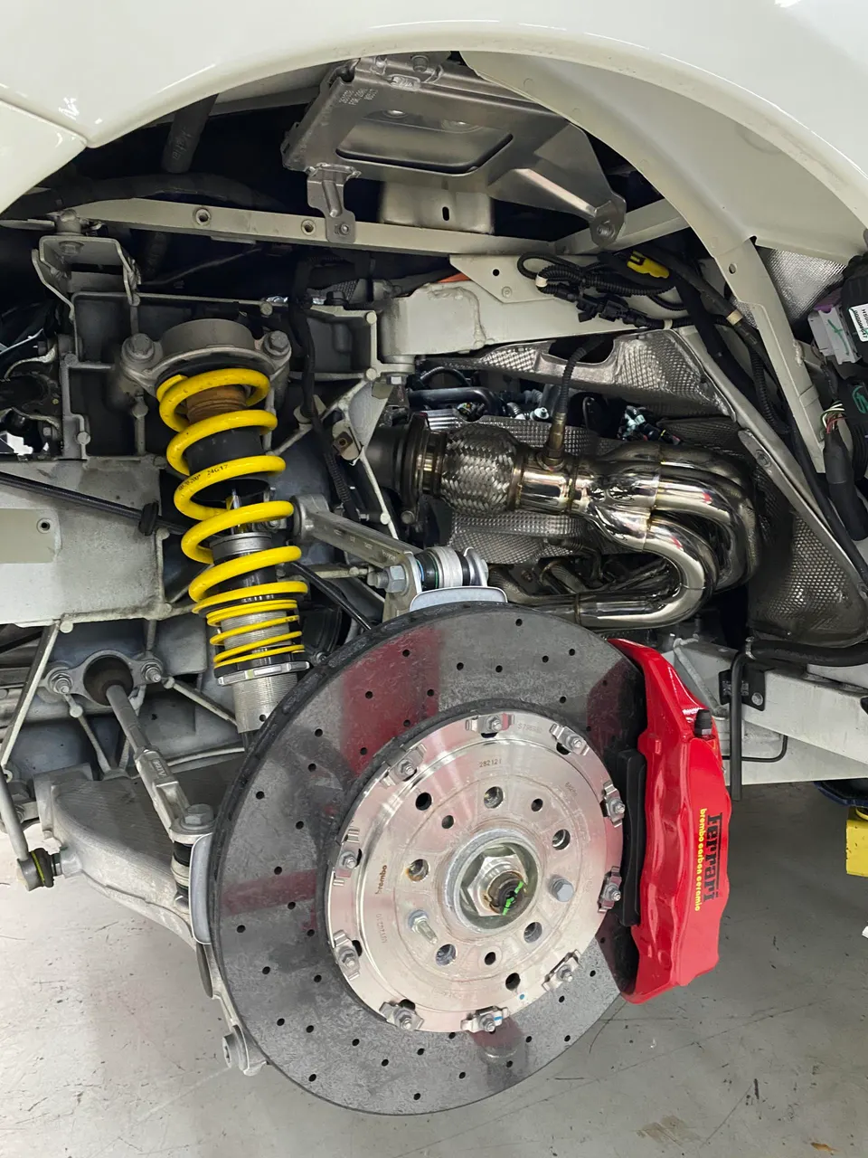 Close-up of a Ferrari's rear suspension, large drilled brake rotor, red Brembo caliper, and performance exhaust headers.