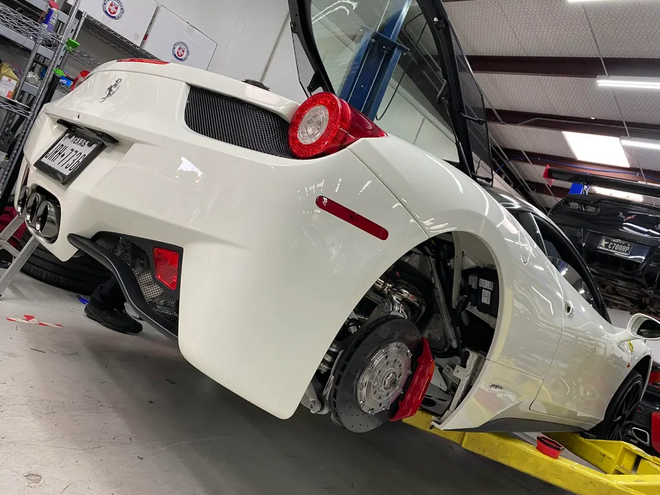 White Ferrari 458 on a lift in a performance shop, with the rear wheel removed exposing red brake calipers and suspension.