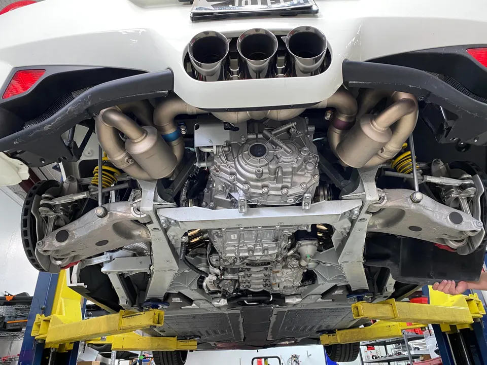 Underside view of a white Porsche 911 on a lift, showcasing its aftermarket exhaust system, transmission, and suspension.