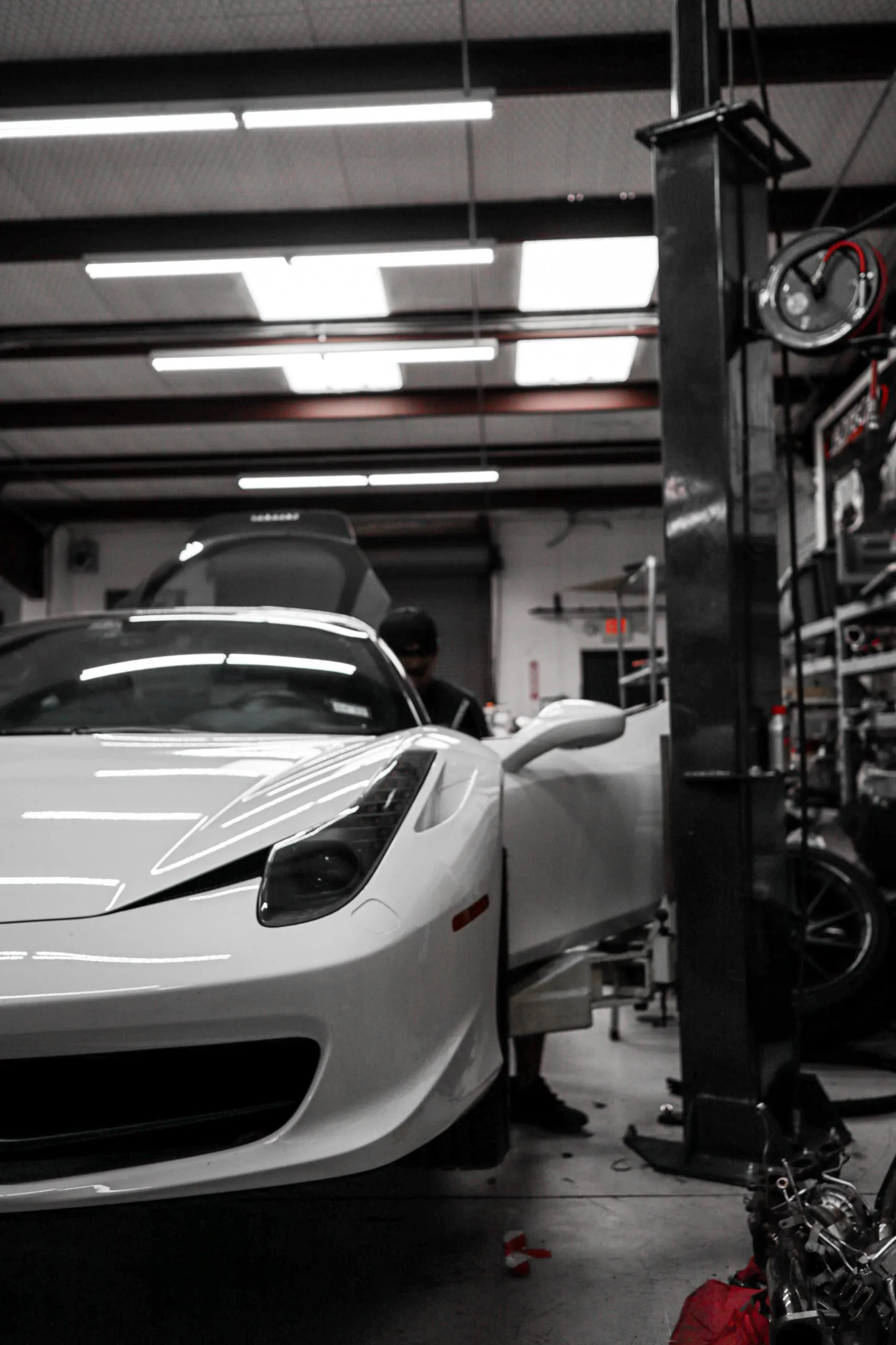A white Ferrari sports car in a performance tuning shop with a mechanic working on it, surrounded by tools and equipment.