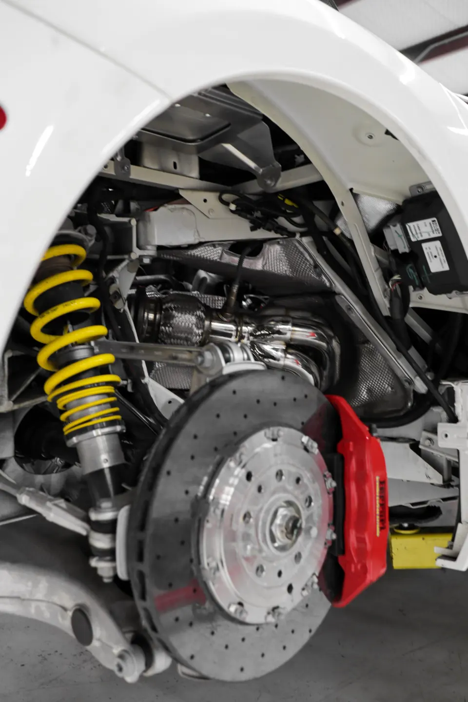Close-up view of a white car's suspension, brake system with red caliper, and exhaust manifold in a shop.