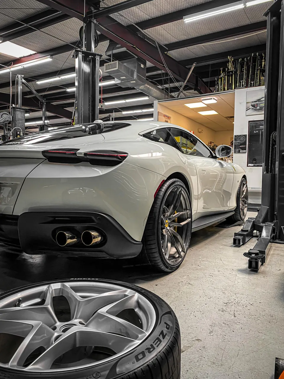 White Ferrari Roma in a performance shop with a wheel removed, showcasing the exhaust and a car lift.