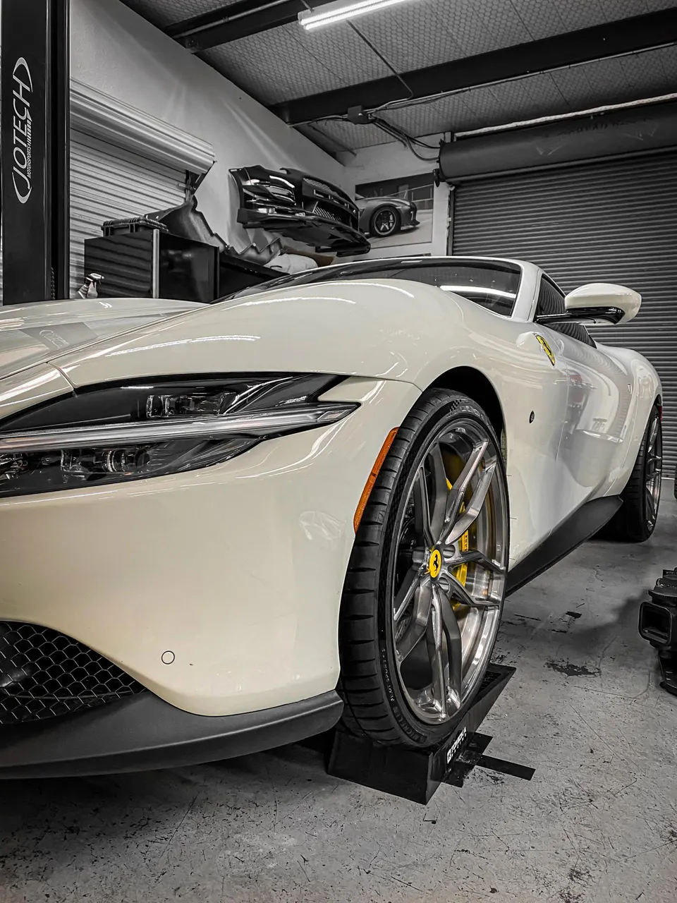 A white Ferrari Roma on ramps in a Jotech Motorsports shop, showcasing its front wheel and yellow brake caliper.