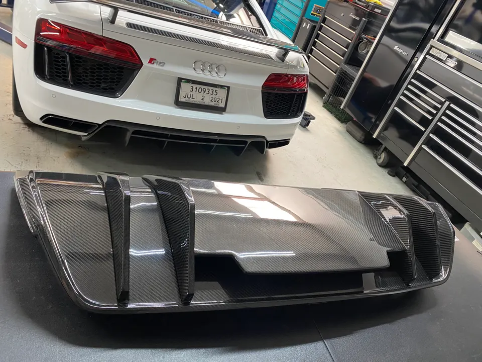 White Audi R8 in a shop with a carbon fiber rear diffuser in the foreground, ready for installation.